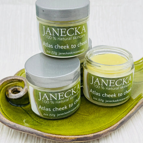Three jars of Janecka Skincare JANECKA Atlas Cheek to Cheek Herbal Healing Ointment are placed on a green ceramic dish, with one jar open to reveal the yellow balm inside.
