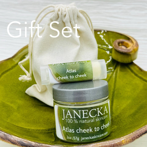 A Janecka Skincare gift set on a green leaf dish includes a small jar of JANECKA Atlas Cheek to Cheek, Herbal Healing Ointment with natural ingredients, plus a drawstring pouch. “Gift Set” text displayed.