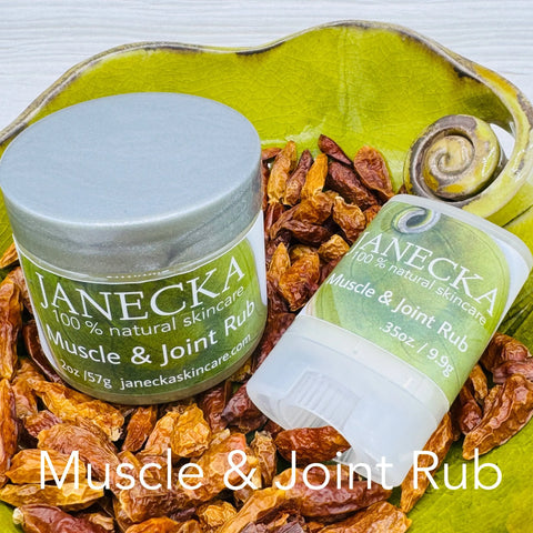 Two Janecka Skincare JANECKA Muscle & Joint Rubs—one jar, one twist-up stick—rest on a green leaf-shaped dish with dried red peppers. “Muscle & Joint Rub” appears at the bottom.