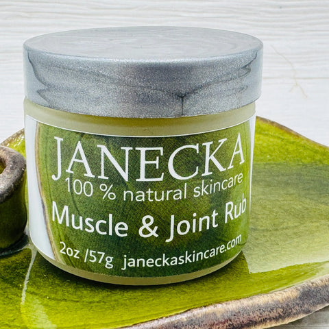 A jar of JANECKA Muscle & Joint Rub by Janecka Skincare sits on a green ceramic tray with a silver lid against a white background, highlighting its herbal pain relief benefits.