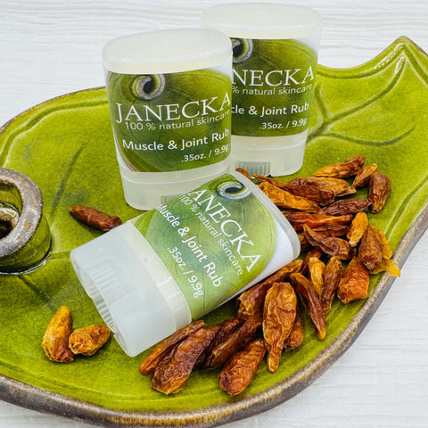 Three JANECKA Muscle & Joint Rub sticks by Janecka Skincare sit on a green ceramic dish with dried chili peppers, showcasing their 100% natural, herbal pain relief and organic skincare benefits.
