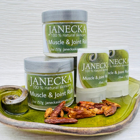 A ceramic plate holds two jars and two sticks of Janecka Skincare JANECKA Muscle & Joint Rub with green labels and silver lids, displayed by dried red chili peppers on a light, wavy-textured background.