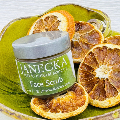 A jar of Janecka Skincare JANECKA Face Scrub sits in a green ceramic dish among dried orange slices. The label is visible, showcasing its gentle exfoliation made with 100% natural ingredients.