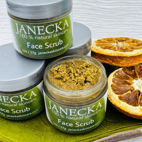 Three JANECKA Face Scrub jars from Janecka Skincare are displayed on a green surface, one open to show the greenish exfoliant. Dried citrus slices beside the jars highlight their natural ingredients.