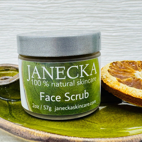 A JANECKA Face Scrub by Janecka Skincare rests on a green ceramic dish beside a dried orange slice, offering gentle exfoliation with natural ingredients against a textured white background.