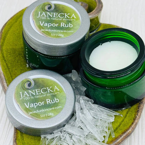 Open green jars of JANECKA Herbal Vapor Rub by Janecka Skincare sit on a green ceramic leaf dish beside scattered menthol crystals. The label reads 100% natural skincare, Herbal Vapor Rub, 1oz / 28g, janeckaskincare.com.