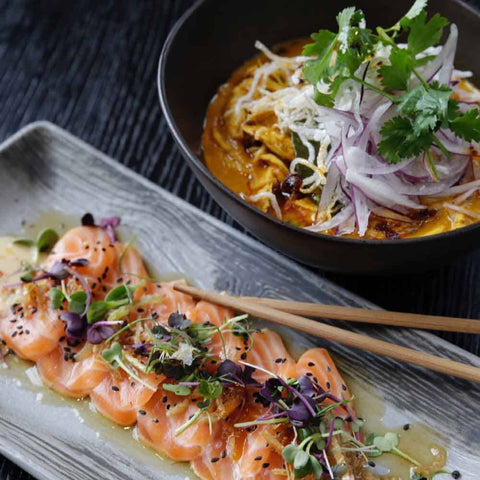 A rectangular plate of salmon sashimi, garnished with microgreens and Gustus Vitae’s Japanese Seasoning Gift Set, sits beside a bowl of noodle soup with red onions and crispy noodles, with chopsticks resting on the plate.