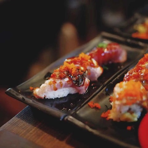 Close-up of sushi topped with fish, seaweed, sauce, and orange roe, elevated by Gustus Vitae’s Japanese Seasoning Gift Set – Tastes of Japan spice blends. Served on a black plate with another sushi piece in the background.