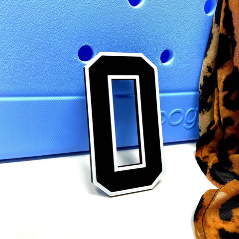 A black and white BOGLETS - Jersey Sports Uniform T-Shirt Number by Chatelet Manufacturing rests against a light blue perforated surface, with part of a brown and black leopard print fabric and a personalized bag charm visible on the right.