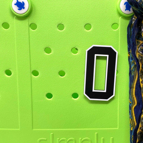 A large black and white BOGLETS - Jersey Sports Uniform T-Shirt Number magnet by Chatelet Manufacturing is used as a personalized bag charm, attached to a bright green surface with round holes, blue turtle magnets, and patterned fabric on the right.