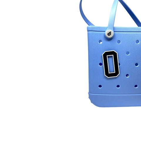 A light blue tote bag with perforated holes, a small button detail by the strap, and a Chatelet Manufacturing BOGLETS Jersey Sports Uniform T-Shirt Number “O” bag charm in black and white attached to the front, set against a white background.