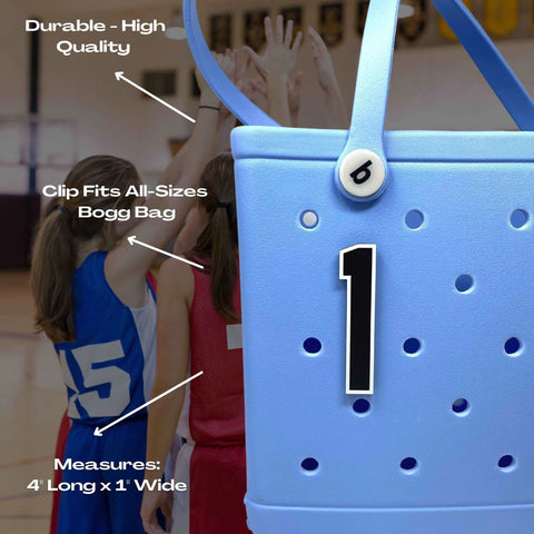 Close-up of a blue Bogg Bag featuring a Chatelet Manufacturing BOGLETS Jersey Sports Uniform T-Shirt Number bag charm; text highlights its durability, adjustable clip, and size. Youth basketball players huddle on the court in the background.