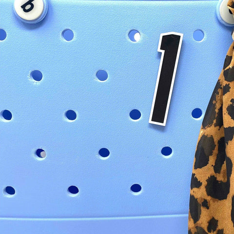 Close-up of the BOGLETS - Jersey Sports Uniform T-Shirt Number by Chatelet Manufacturing, shown on a light blue perforated surface with letter beads and leopard-print strap—ideal as sports bag charms or tote bag accessories.