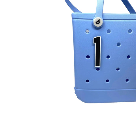 A close-up of a light blue rubber tote bag featuring a Chatelet Manufacturing BOGLETS - Jersey Sports Uniform T-Shirt Number charm in black-and-white. Only part of the bag and one handle are shown, highlighting this personalized accessory.