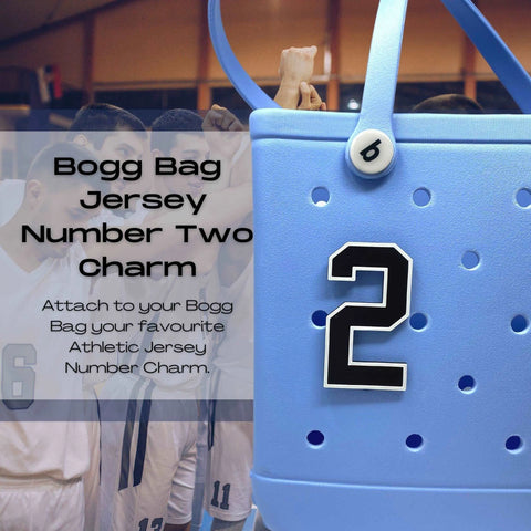 A blue Bogg Bag features a Chatelet Manufacturing BOGLETS Jersey Sports Uniform T-Shirt Number bag charm in black and white. In the background, a basketball team huddles, highlighting this fun tote accessory for sports fans.