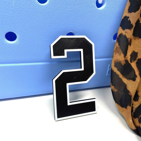 The BOGLETS - Jersey Sports Uniform T-Shirt Number by Chatelet Manufacturing, in black and white plastic, stands before a light blue perforated board beside leopard print fabric—ideal as a personalized bag charm or tote accessory.