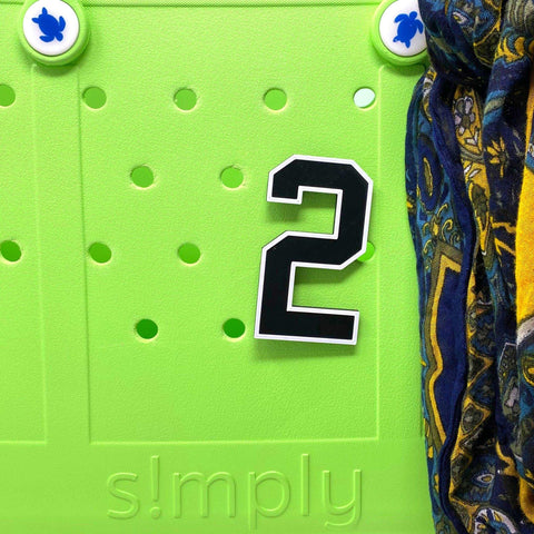 The BOGLETS - Jersey Sports Uniform T-Shirt Number by Chatelet Manufacturing is a large black and white 2 magnet, perfect as a personalized bag charm, displayed on a green board with blue turtle knobs and patterned fabric on the right.