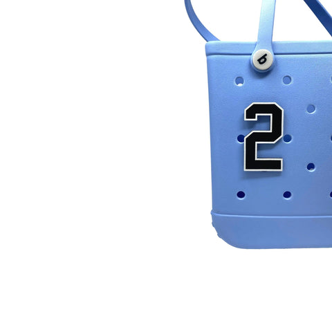 A partial view of a light blue tote bag with round holes features a Chatelet Manufacturing BOGLETS Jersey Sports Uniform T-Shirt Number (black-and-white “2”) bag charm. The bag has handles, a top button detail, and is perfect for displaying accessories. White background.