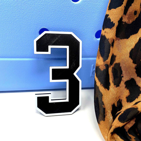A black and white BOGLETS Jersey Sports Uniform T-Shirt Number by Chatelet Manufacturing stands on a white surface, with a blue perforated background and leopard print fabric, ideal for displaying personalized bag charms or tote accessories.