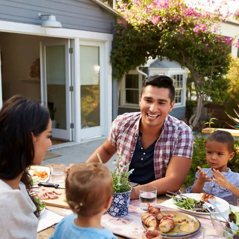 A family of four enjoys a garden meal by their house, savoring dishes enhanced with Gustus Vitae Kid’s Menu: Italian: Pasta & Pizza Seasoning, as two adults and two kids smile, chat, and appreciate the tasty, kid-friendly flavors outdoors.