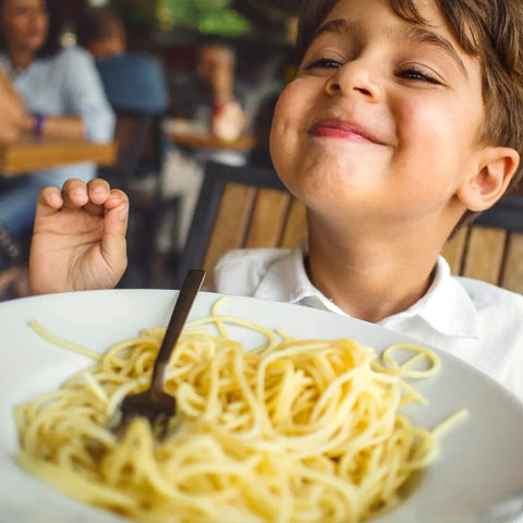 A smiling child sits at a table in a restaurant, excitedly looking at a plate of spaghetti topped with Gustus Vitae Kid’s Menu: Italian: Pasta & Pizza Seasoning, with a fork ready in front of them.