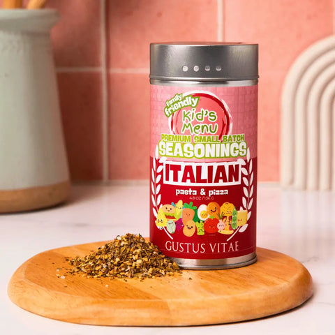 A container of Gustus Vitae Kid’s Menu: Italian: Pasta & Pizza Seasoning sits on a wooden board beside a small pile of the seasoning, with kitchen utensils and a pink tiled wall in the background.