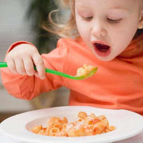 A young child in an orange shirt holds a green fork, ready to taste pasta topped with Gustus Vitae Kid’s Menu: Italian: Pasta & Pizza Seasoning from a white plate, gazing eagerly at the food with their mouth open.