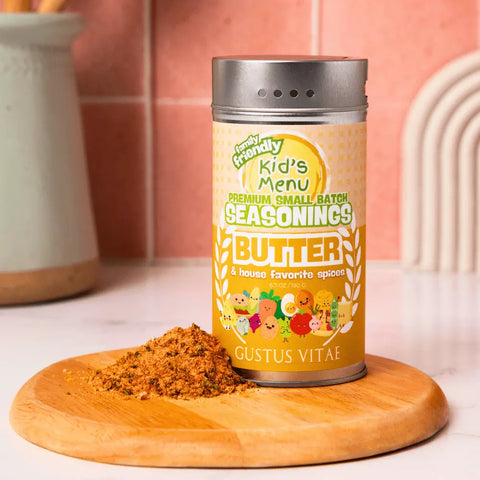 A container of Gustus Vitae Kid’s Menu: Butter and House Favorite Spices sits on a wooden board beside a small pile of this gourmet seasoning, with a kitchen backdrop featuring pink tiles and a utensil holder.