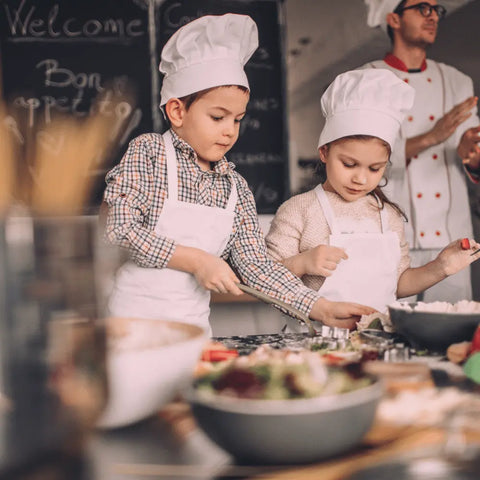 Two young children in chef hats and aprons mix ingredients with Gustus Vitae’s Kid’s Menu: Butter: And House Favorite Spices at a kitchen counter, as a chef and chalkboard menu appear in the background.
