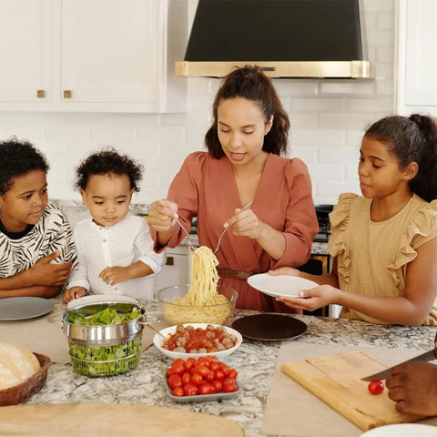 A woman serves spaghetti to a child as three kids watch at a kitchen counter filled with salad, tomatoes, and meatballs seasoned with Gustus Vitae’s Kid’s Menu: Butter & House Favorite Spices. The scene is warm and lively.