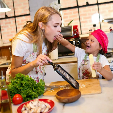 A young woman and smiling child in aprons cook with Gustus Vitae’s Kid’s Menu: Butter & House Favorite Spices, as the child feeds her cheese and holds a grater amid fresh veggies on the kitchen counter.