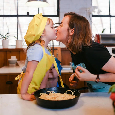 A young child and an adult in aprons and chef hats share a kiss in the kitchen, while a skillet of spaghetti made with Gustus Vitaes Kid’s Menu: Butter: And House Favorite Spices sits on the counter. Another child peeks from behind.