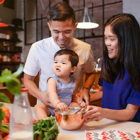A smiling family gathers in the kitchen as an adult helps a baby mix fresh ingredients using Gustus Vitaes Kid’s Menu: Butter & House Favorite Spices, with fresh vegetables and a bottle on the counter.