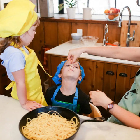 Two kids in colorful aprons and chef hats are in the kitchen. An adult feeds spaghetti, made with Gustus Vitae Kid’s Menu: Butter & House Favorite Spices, to one child while the other watches. A skillet of spaghetti sits on the counter.