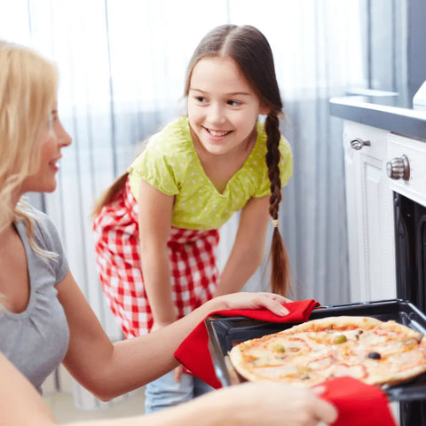 A woman uses oven mitts to take a pizza seasoned with Gustus Vitae Kid’s Menu: Butter & House Favorite Spices from the oven, as a smiling young girl in a green shirt with braids watches her in the kitchen.