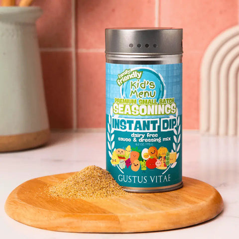 A container of Gustus Vitae Kid’s Menu: Instant Dip: Dairy-Free Sauce & Dressing Mix sits on a wooden board with some mix beside it; kitchen utensils and a pink tiled wall are in the background, highlighting this tasty, dairy-free dip.