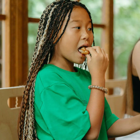 A young girl in a green shirt enjoys food with Gustus Vitae Kid’s Menu: Instant Dip, a dairy-free sauce & dressing mix, while sitting indoors near a window with greenery outside.