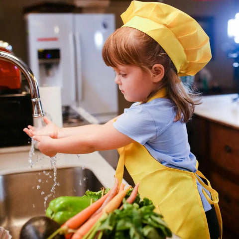 A young child in a yellow chef hat and apron washes up at the sink. Fresh carrots and bell peppers sit on the counter next to Gustus Vitae’s Kid’s Menu: Instant Dip—Dairy-Free Sauce & Dressing Mix, ready for a quick snack.