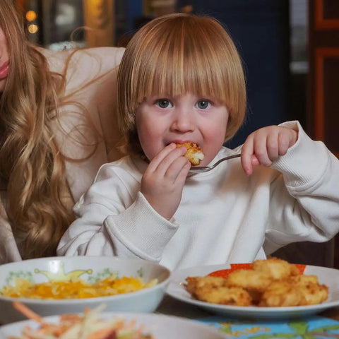 A blonde child in a white shirt enjoys food while holding a fork at a table set with plates featuring Gustus Vitae’s Kid’s Menu: Instant Dip: Dairy-Free Sauce & Dressing Mix. An adult with long hair sits partially visible beside them.