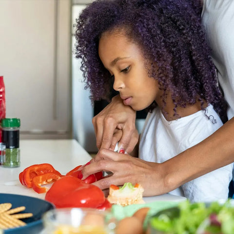 A curly-haired child slices a red bell pepper on the counter, guided by an adult, with fresh vegetables and Gustus Vitae’s Kid’s Menu: Instant Dip—Dairy-Free Sauce & Dressing Mix visible nearby.