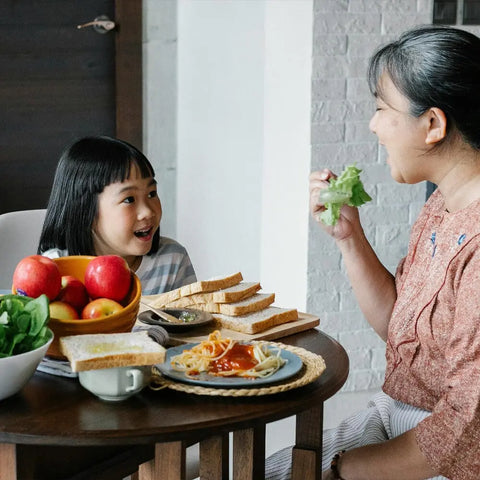 A woman and a young girl smile at each other while sharing a meal in a bright, cozy room, enjoying fresh food enhanced by Gustus Vitae Kid’s Menu: Instant Dip—an easy, dairy-free sauce & dressing mix that adds extra flavor to their spread.