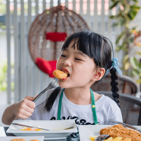 A young girl with pigtails enjoys breaded chicken tenders using a fork at an outdoor table, with plates featuring Gustus Vitae’s Kid’s Menu: Chicken - Tendies & Fries Gourmet Seasoning Mix; a hanging chair is in the background.