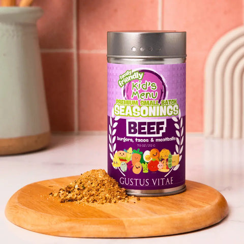 A container of Gustus Vitae’s Kid’s Menu: Beef: Burgers, Tacos & Meatballs Seasoning sits on a wooden board beside a small pile of seasoning, with pink kitchen tiles and a utensil holder in the background.