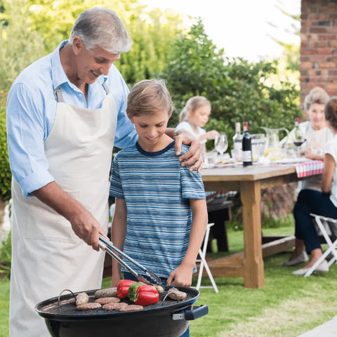 An older man in an apron grills sausages and bell peppers with a young boy beside him, using Gustus Vitae’s Kid’s Menu: Beef: Burgers, Tacos & Meatballs Seasoning, while two women enjoy an outdoor meal at a picnic table in the background.