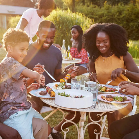 A family enjoys an outdoor meal, smiling as they share salads and skewers at a sunny garden table. Gustus Vitae Kid’s Menu: Beef: Burgers, Tacos & Meatballs Seasoning brings kid-friendly flavor to their feast while others chat nearby.