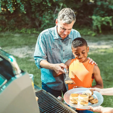 A man grills burgers outdoors, placing a patty seasoned with Gustus Vitae Kid’s Menu: Beef: Burgers, Tacos & Meatballs Seasoning onto a plate held by a smiling boy in an orange shirt as they enjoy a sunny backyard day.