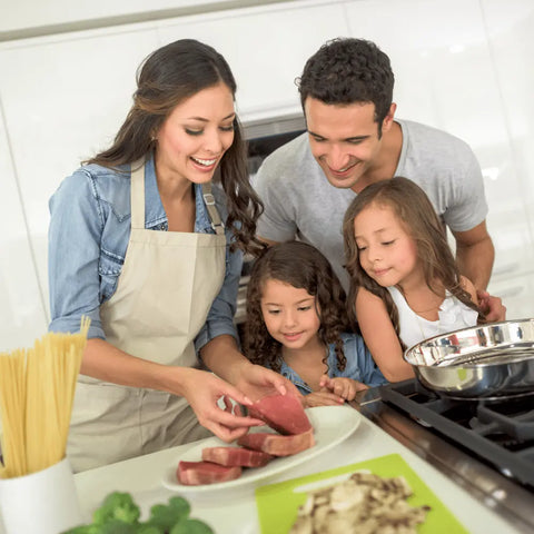 A smiling family of four prepares a meal in a bright kitchen, as the mother seasons raw steaks with Gustus Vitae Kid’s Menu: Beef: Burgers, Tacos & Meatballs Seasoning while her two daughters watch and the father stands nearby at the stove.