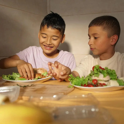 Two boys enjoy sandwiches with lettuce and tomatoes, smiling in warm natural light—thanks to Gustus Vitae’s Kid’s Menu: Veggie: All Purpose Seasoning, a kid-approved blend made with non-GMO ingredients that adds extra flavor to every bite.