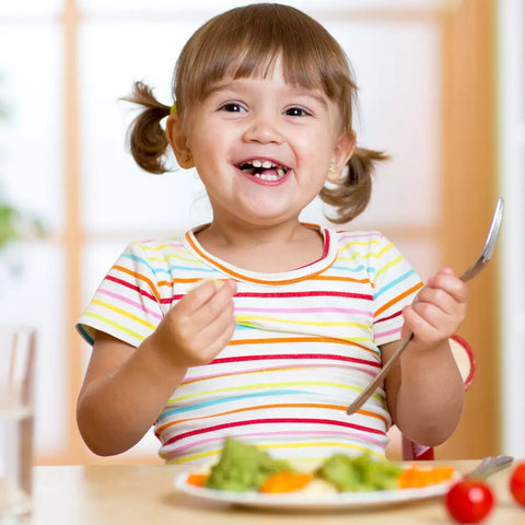 A smiling young girl in a striped shirt sits at the table, happily enjoying veggies seasoned with Gustus Vitae Kid’s Menu: Veggie: All Purpose Seasoning, looking joyful about her tasty and healthy meal.