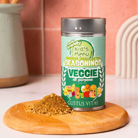 A green Gustus Vitae Kid’s Menu: Veggie: All Purpose Seasoning canister sits on a wooden board beside a small pile of this non-GMO, kid-approved seasoning, with a light countertop and blurred kitchen items in the background.